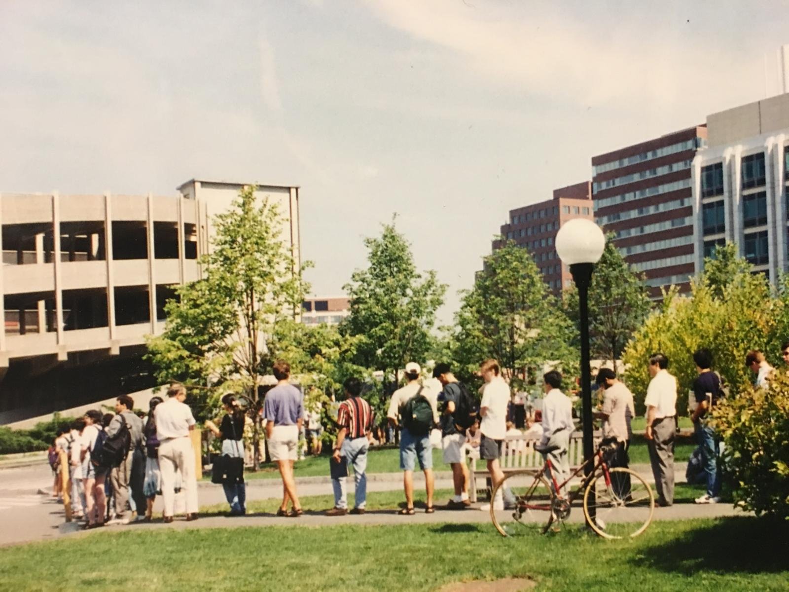 A lunchtime line of students and staff stretching along the path at MIT on a sunny day, seen from the green, past a bicycle leaning against a lamp post.