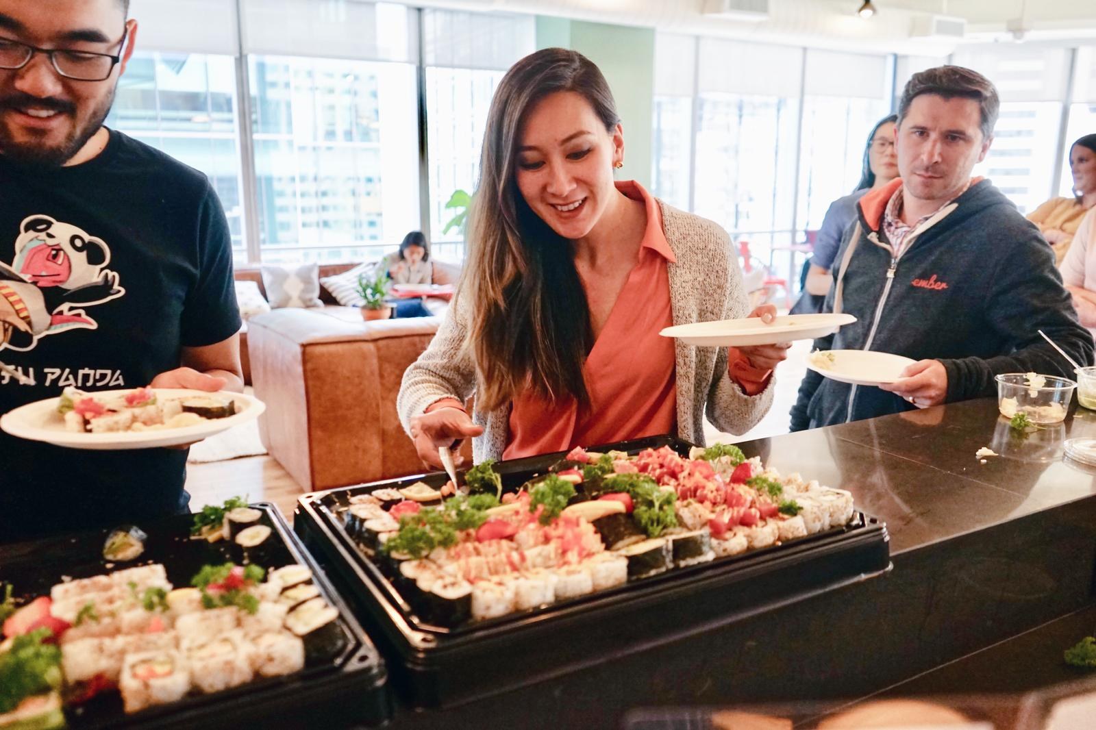 Three colleagues filling their plates from sushi platters at an office counter, city windows behind them.