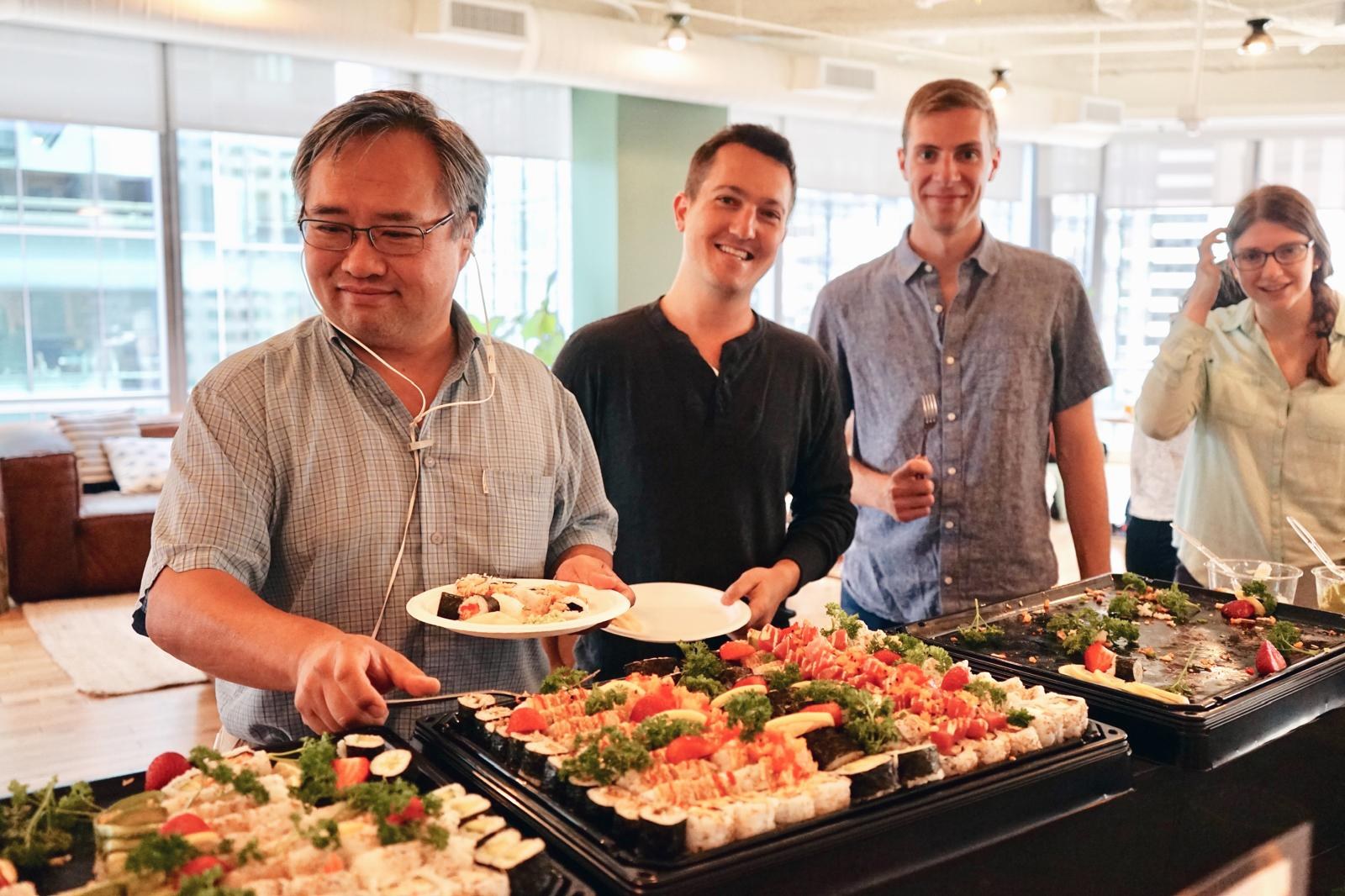 Three men holding plates beside a sushi spread in a bright office.