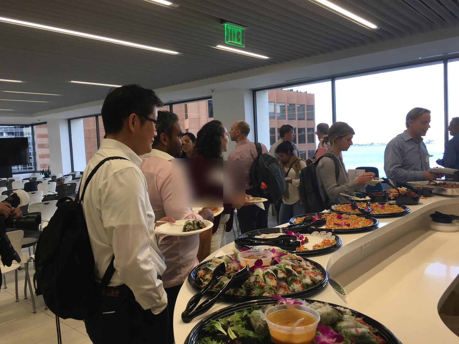 A lunch line of office workers by a wall of windows, plates in hand.