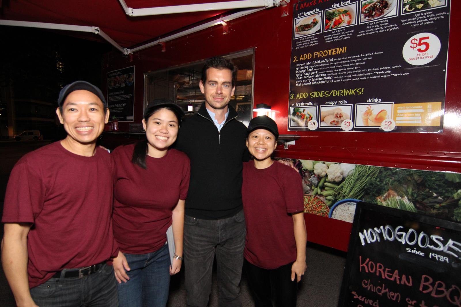 Three of the crew in maroon shirts standing with a customer at the lit serving window of the MomoGoose truck at night. A chalkboard beside them reads MomoGoose, since 1989.
