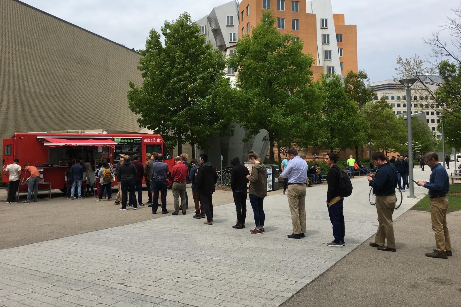 A long midday line leading to a red food truck with a striped awning outside MIT's Stata Center, many people in the queue looking at their phones.